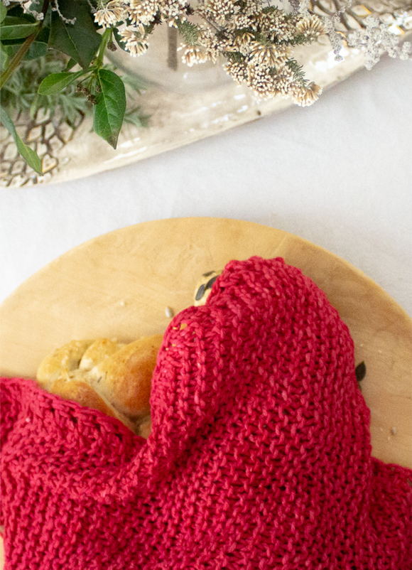 Red handmade chunky-knit Penelope D challah cover over a challah on a wooden board with table setting